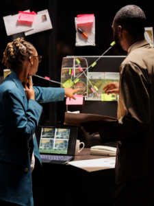 Two people stand in a dimly lit workspace analyzing a board covered with photos, notes, and string connections. One person points toward an image on the board while the other holds a folder. A laptop displaying surveillance-style images, a coffee mug, and a desk phone sit on a table nearby, suggesting an investigative or analytical setting.