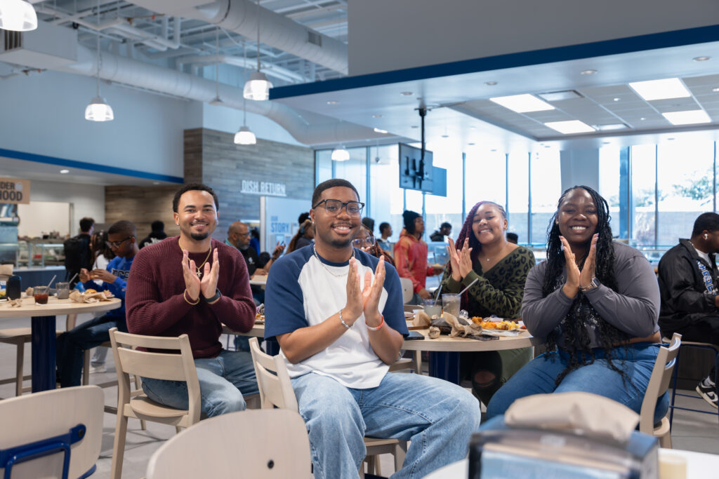 Four students sit at a table inside a campus dining hall, clapping while facing the camera. The space features bright lighting, large windows, and overhead fixtures, with other students eating and socializing in the background near food stations labeled “Dish Return.”