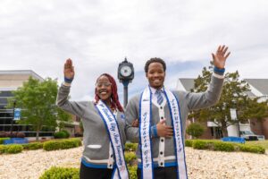 Mister and Miss ECSU waving while interlocking arms. Standing outside in front of the clock tower.