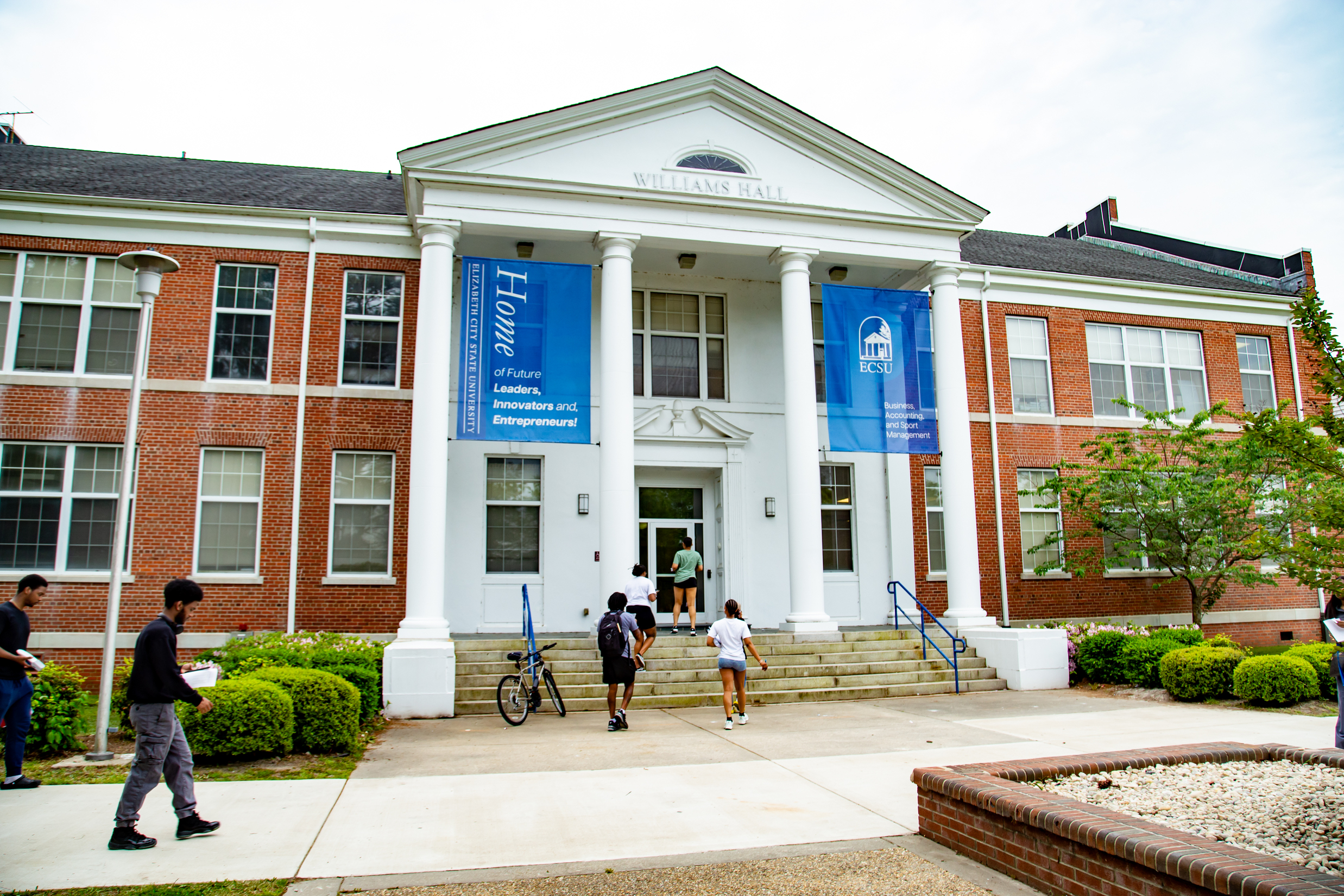 Front entrance of Williams Hall at ECSU, a red-brick academic building with white columns and a triangular pediment labeled “Williams Hall.” Students walk up the steps and enter the building, with blue ECSU banners hanging between the columns.