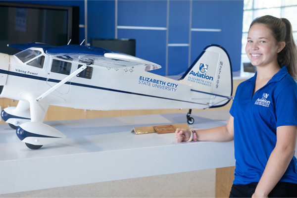 ECSU aviation student stands beside a detailed model aircraft labeled “Elizabeth City State University Aviation” on display indoors, smiling in a modern academic space with blue walls and presentation screens.