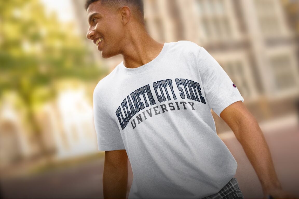 Student wearing a white “Elizabeth City State University” T‑shirt smiles and looks to the side while walking outdoors in a sunlit campus setting with blurred buildings and trees in the background.