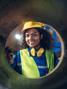 A person wearing a yellow hard hat, high-visibility safety vest, and protective earmuffs stands inside a large metal pipe or cylindrical structure. The person holds a handheld inspection device and faces the camera. Industrial lighting and equipment are visible in the background, suggesting a factory or work site setting.