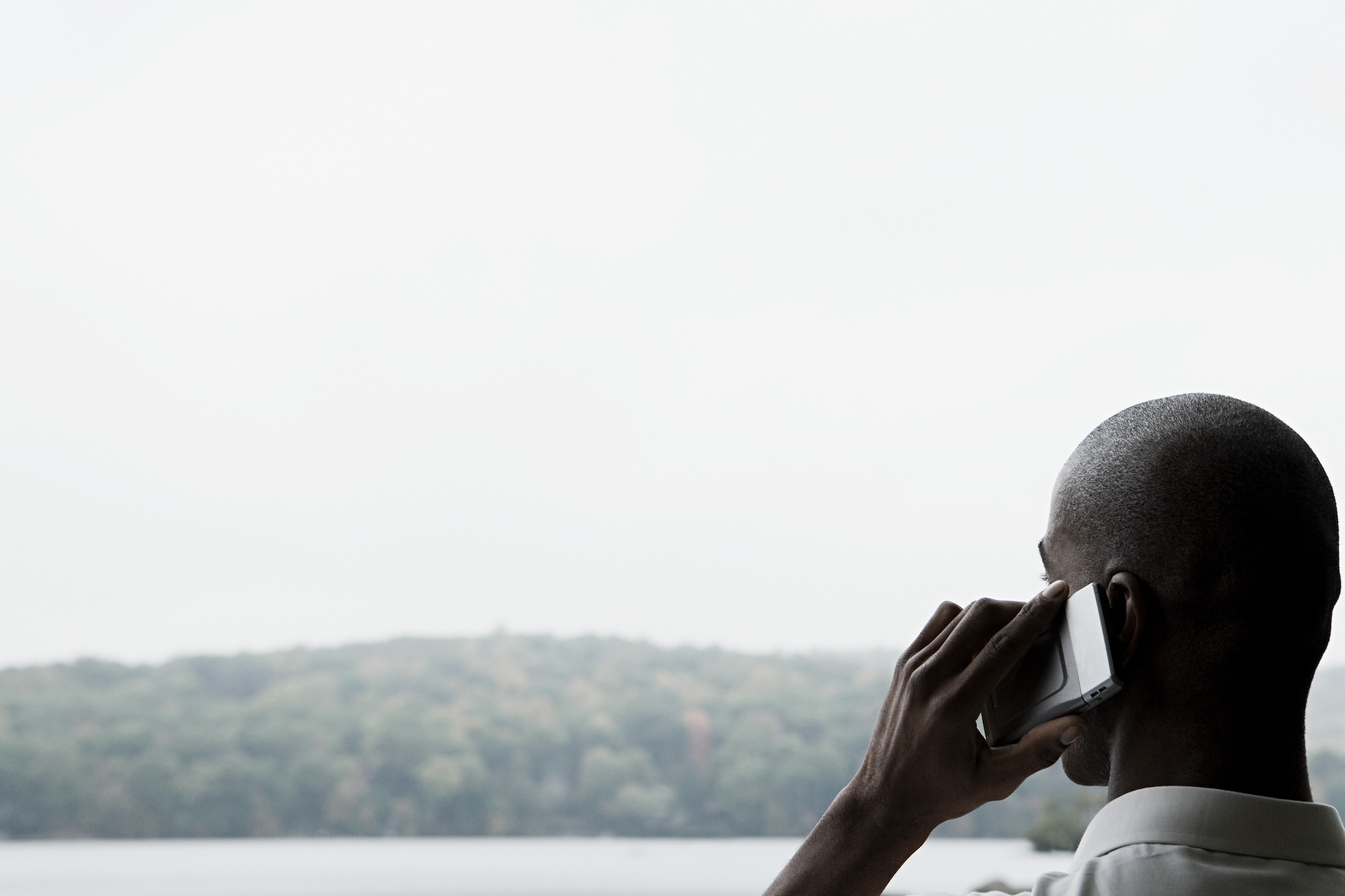 A man is having a phone conversation as he looks out over a lake and forested landscape.