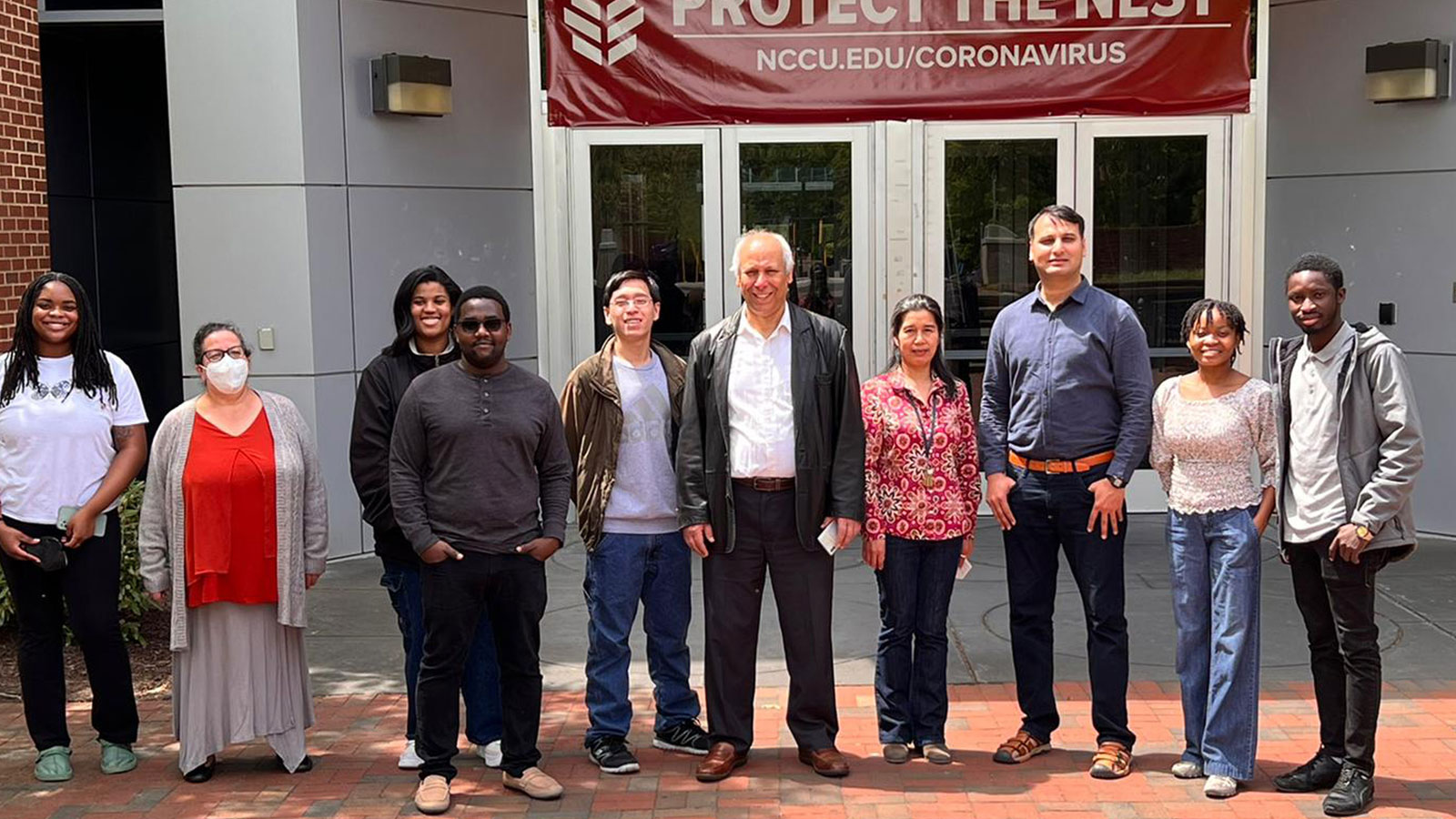 Group of students and faculty stand together outdoors in front of a building entrance, beneath a banner that reads “Protect the Nest” with a website link to NCCU coronavirus information. The group faces the camera and poses for a photo on a brick walkway.