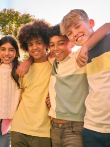 Four school-age children stand close together outdoors with their arms around each other’s shoulders, facing the camera. The group wears casual clothing in light colors, and sunlight creates a warm glow. Trees and bright sky are visible in the background, suggesting a park or outdoor setting.