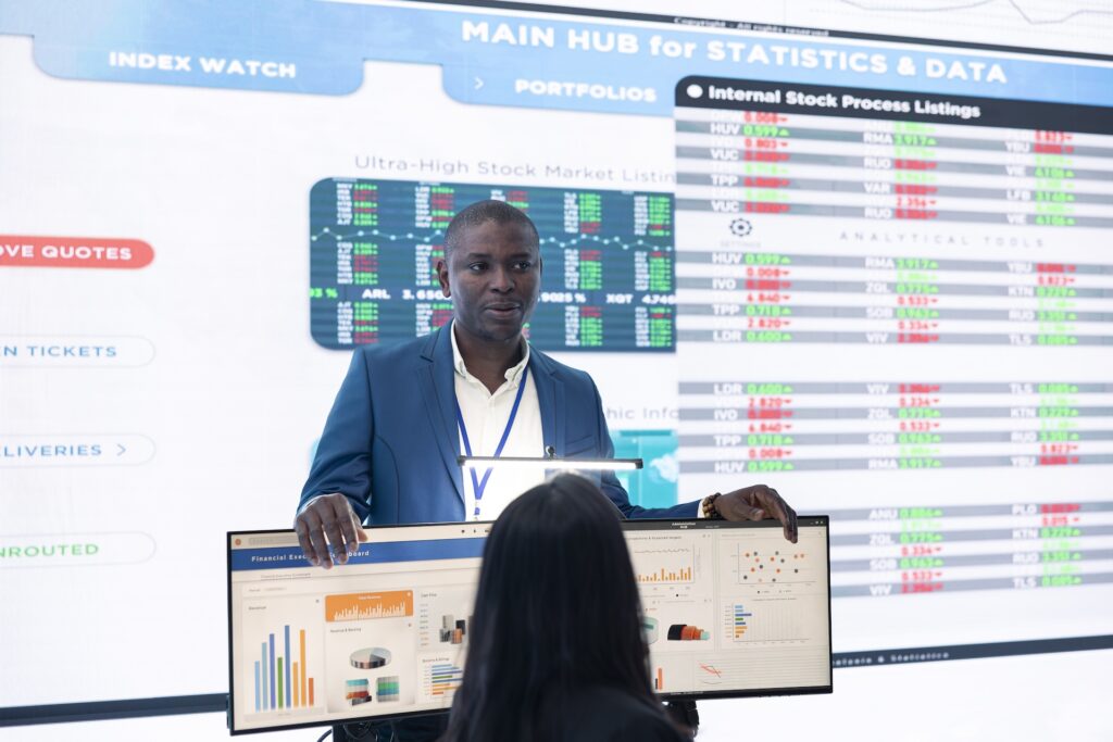 A person with short dark hair in a blue suit gestures towards a flat screen monitor that displays financial data.