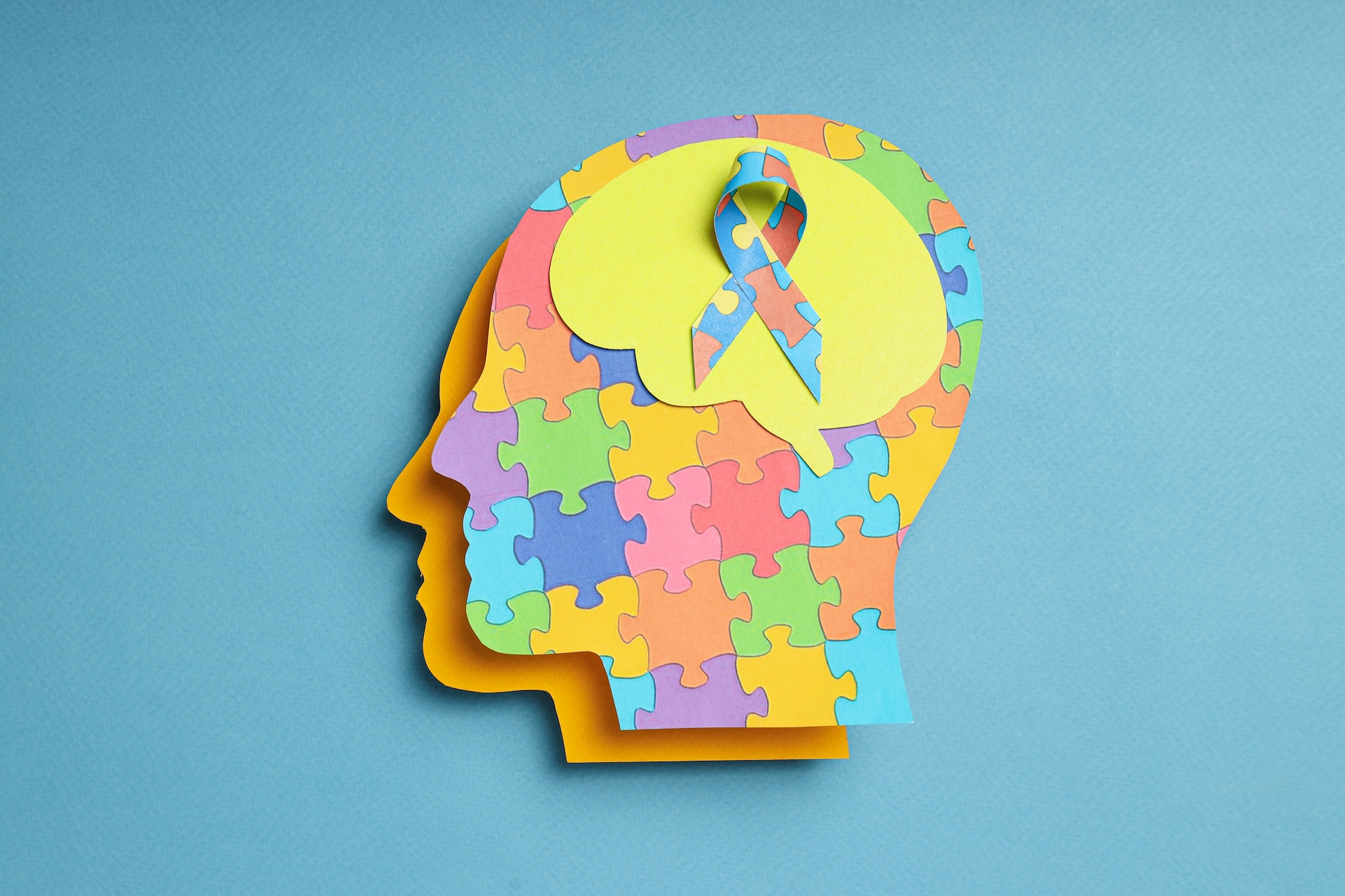 An abstract paper cutout of a head is constructed of puzzle pieces, with a yellow paper cutout of a brain containing a ribbon made from puzzle pieces. 