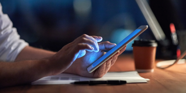 Close-up of a person using a tablet at a desk, with the screen glowing in a dimly lit environment. A coffee cup and papers sit nearby, suggesting studying, work, or accessing online resources.