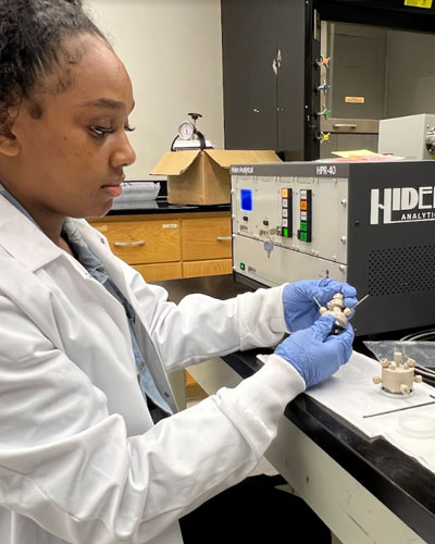 Student wearing gloves works with a small component in a laboratory while operating a scientific instrument, focusing on a detailed hands-on research task.