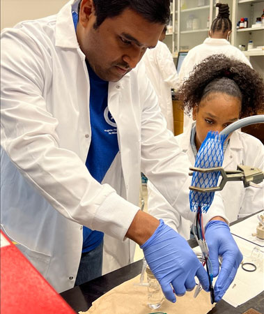 Instructor and student work together in a laboratory, both wearing lab coats and gloves, as they handle equipment and materials during a hands-on experiment.