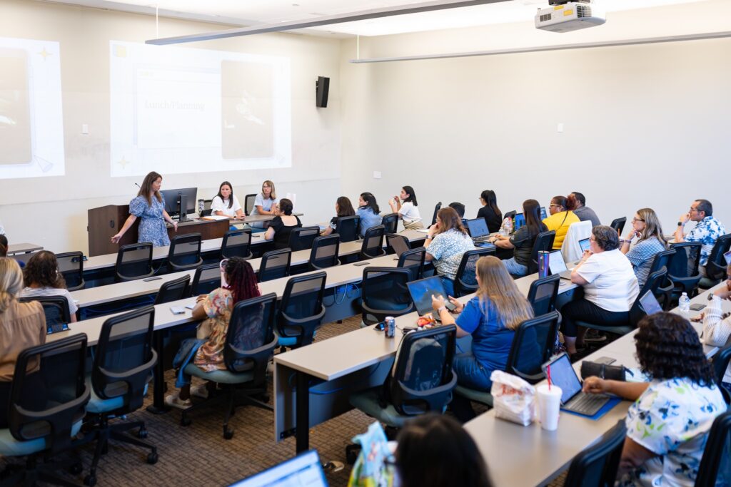 Instructor leads a session in a large classroom while participants sit at tiered desks using laptops and listening to a presentation displayed on a screen.
