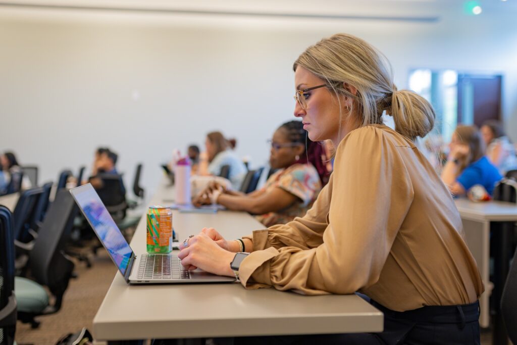 Student works on a laptop in a classroom while other students sit at desks behind her, creating a focused learning environment.