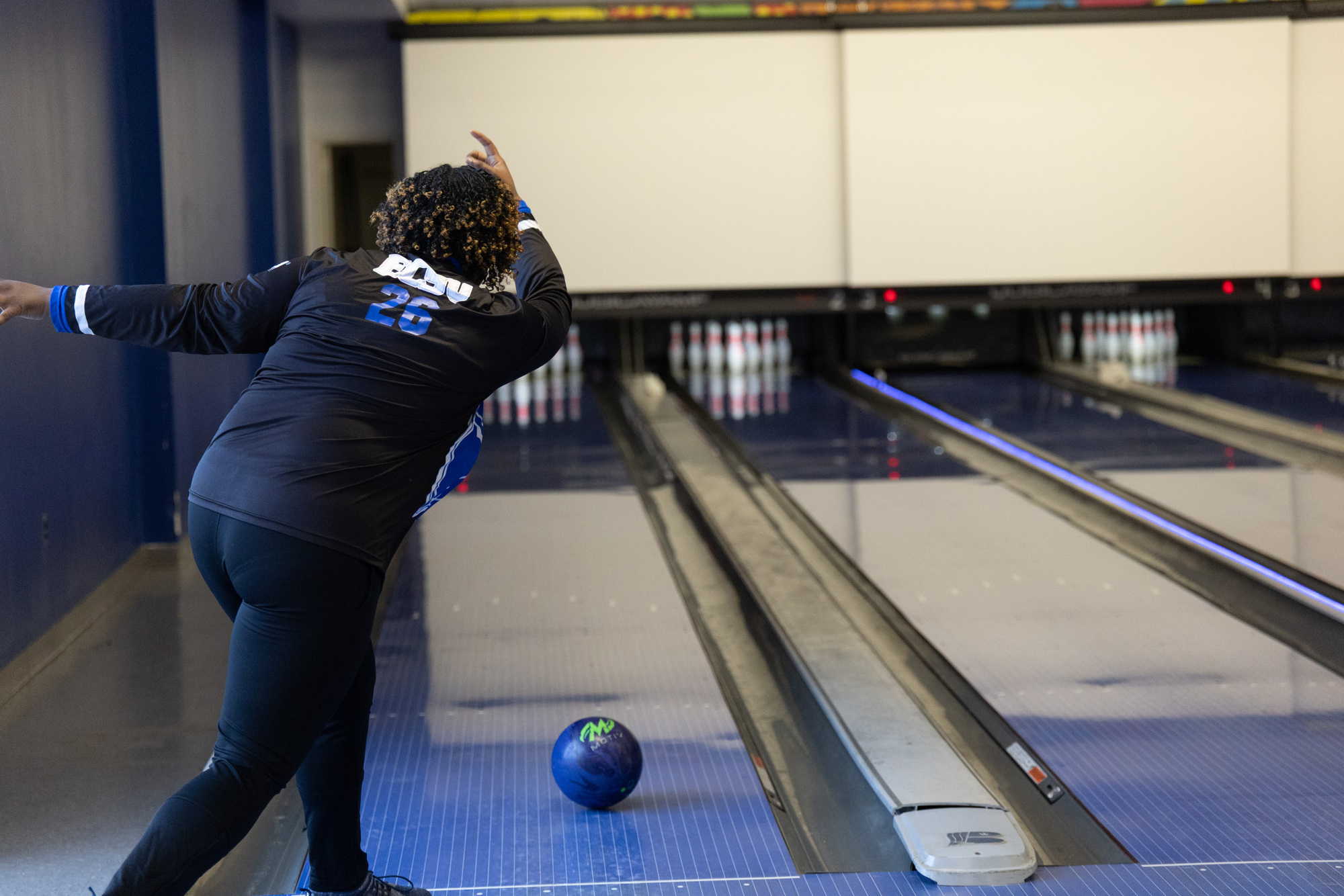 Student bowls on an indoor bowling lane, releasing a blue bowling ball toward the pins while wearing a black and blue jersey.