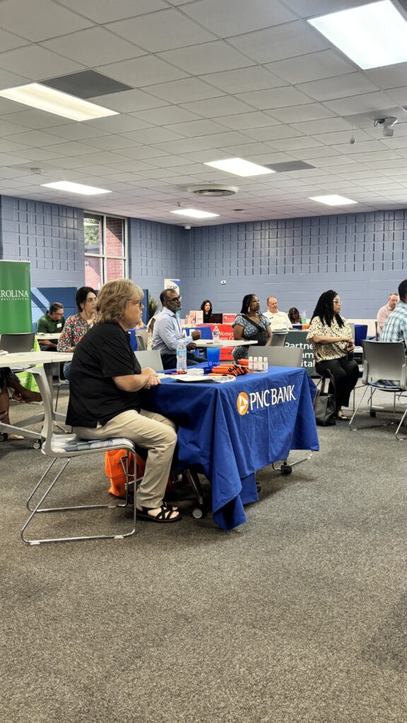 Attendees sit at tables and listen during a small business networking and educational event. Representatives from organizations including PNC Bank and other community partners are seated throughout the room.