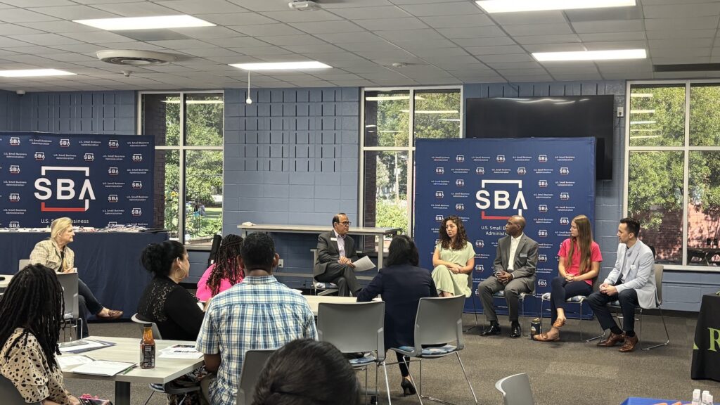Panelists participate in a small business discussion during a U.S. Small Business Administration event while attendees listen from tables in a conference room. SBA-branded displays are visible behind the speakers.