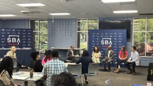 Panelists participate in a small business discussion during a U.S. Small Business Administration event while attendees listen from tables in a conference room. SBA-branded displays are visible behind the speakers.