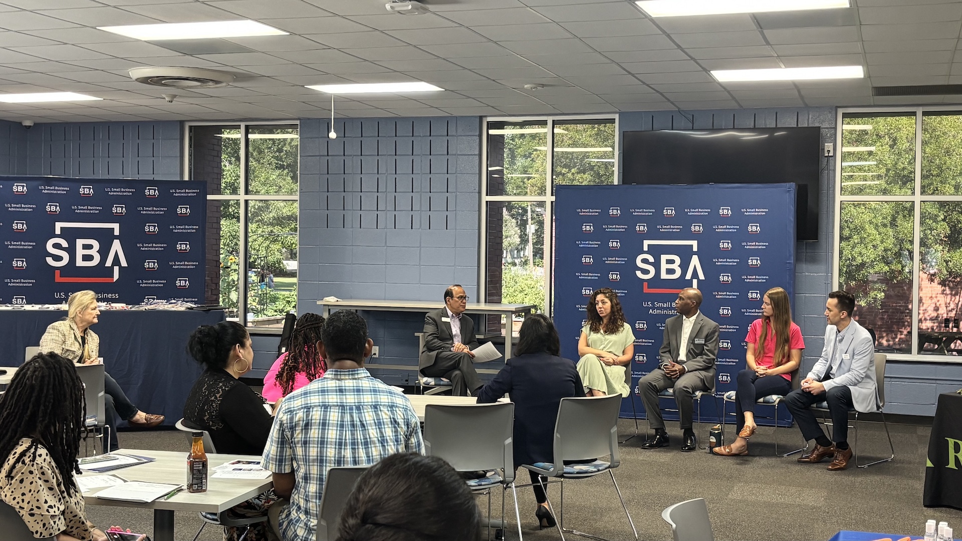 Panelists participate in a small business discussion during a U.S. Small Business Administration event while attendees listen from tables in a conference room. SBA-branded displays are visible behind the speakers.