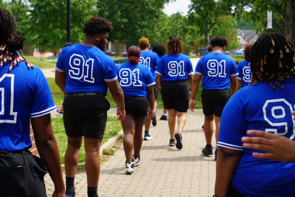 Group of ECSU students wearing matching blue shirts with the number “91” walk together along a campus pathway, viewed from behind, with trees and buildings in the background.