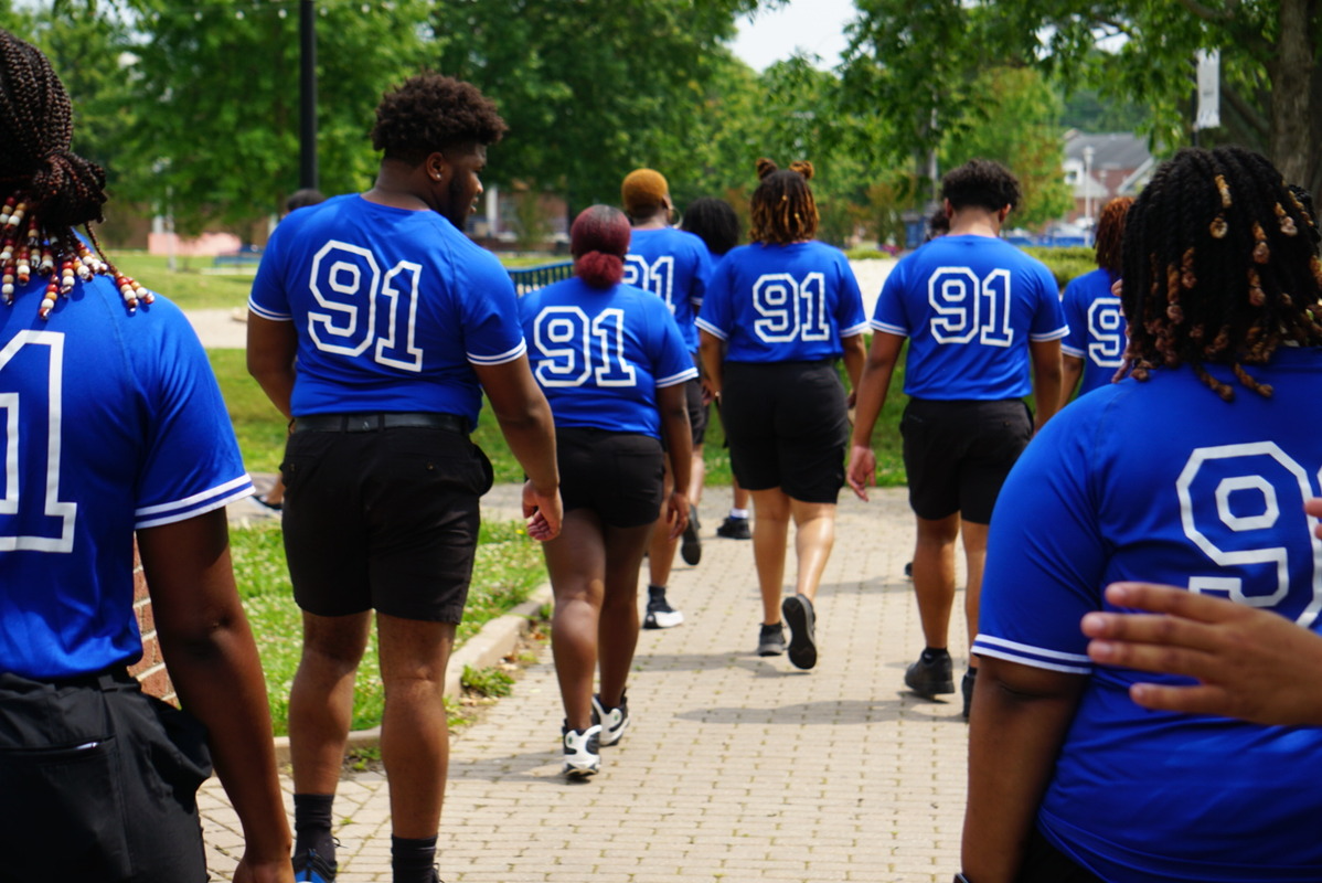 Group of ECSU students wearing matching blue shirts with the number “91” walk together along a campus pathway, viewed from behind, with trees and buildings in the background.