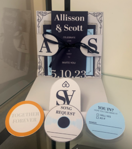 Wedding invitation suite inspired by music and vinyl records, featuring a layered invitation package tied with a black ribbon, custom RSVP cards, song request cards designed to resemble compact discs, and coordinating inserts in navy, white, and light blue.