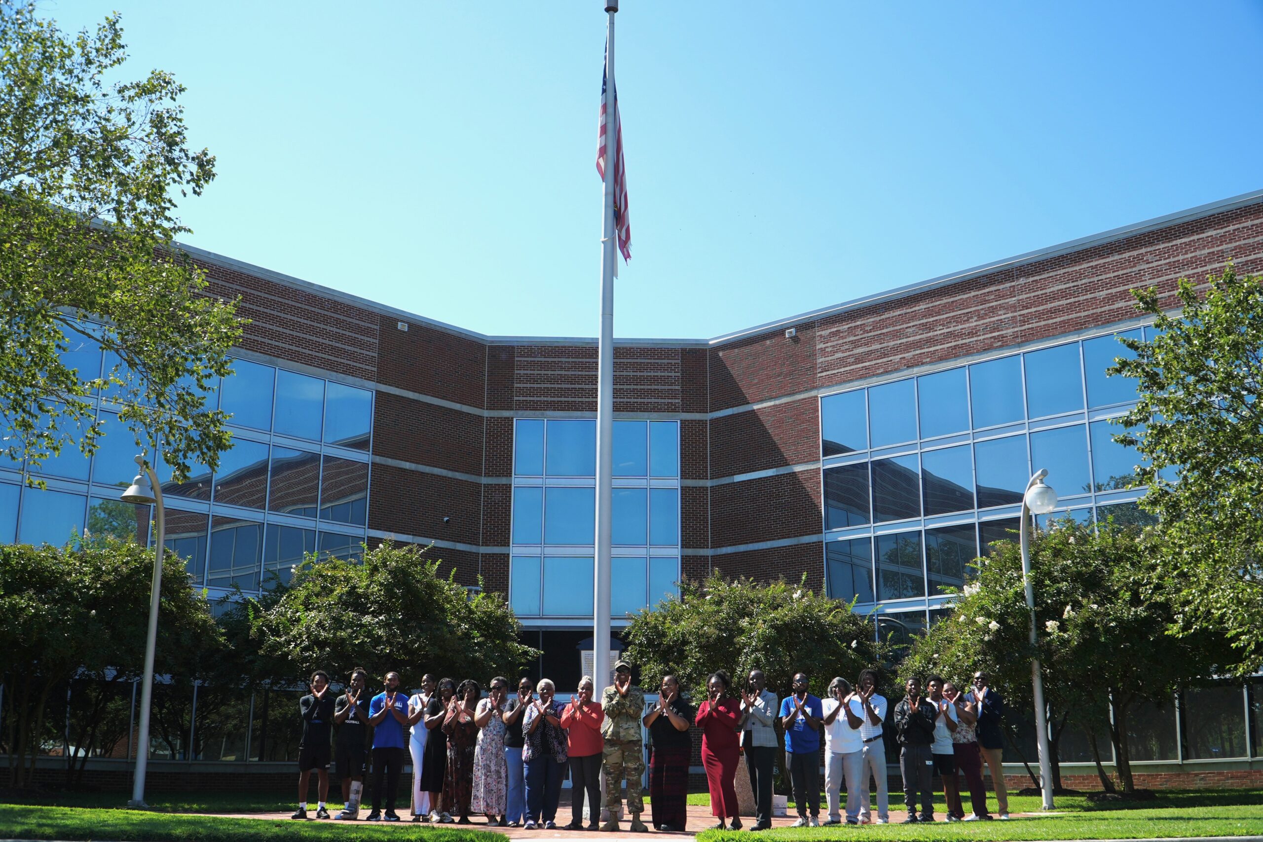 Large group of ECSU students, faculty, staff, and military personnel stand together outdoors in front of a modern campus building with large glass windows and a flagpole. The group is gathered on a walkway and lawn area beneath the American flag, posing for a campus photo.