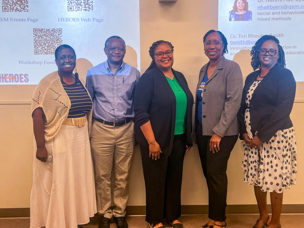 Five faculty and research professionals pose for a photo in front of a presentation screen displaying information about the HEROES research program, including QR codes and workshop resources.