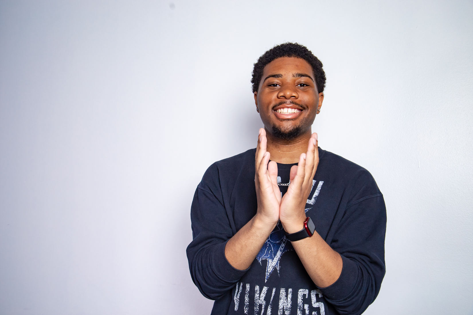 ECSU student stands against a light-colored wall, wearing a black Vikings sweatshirt and smartwatch. The student faces the camera with hands raised together in front of the chest.