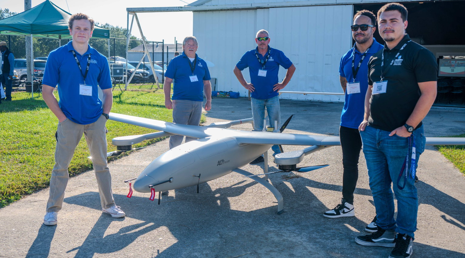 ECSU Aviation students and faculty stand outdoors around a large unmanned aircraft system (UAS) on display near a hangar. The drone features multiple rotors and fixed wings, showcasing advanced aviation and drone technology.